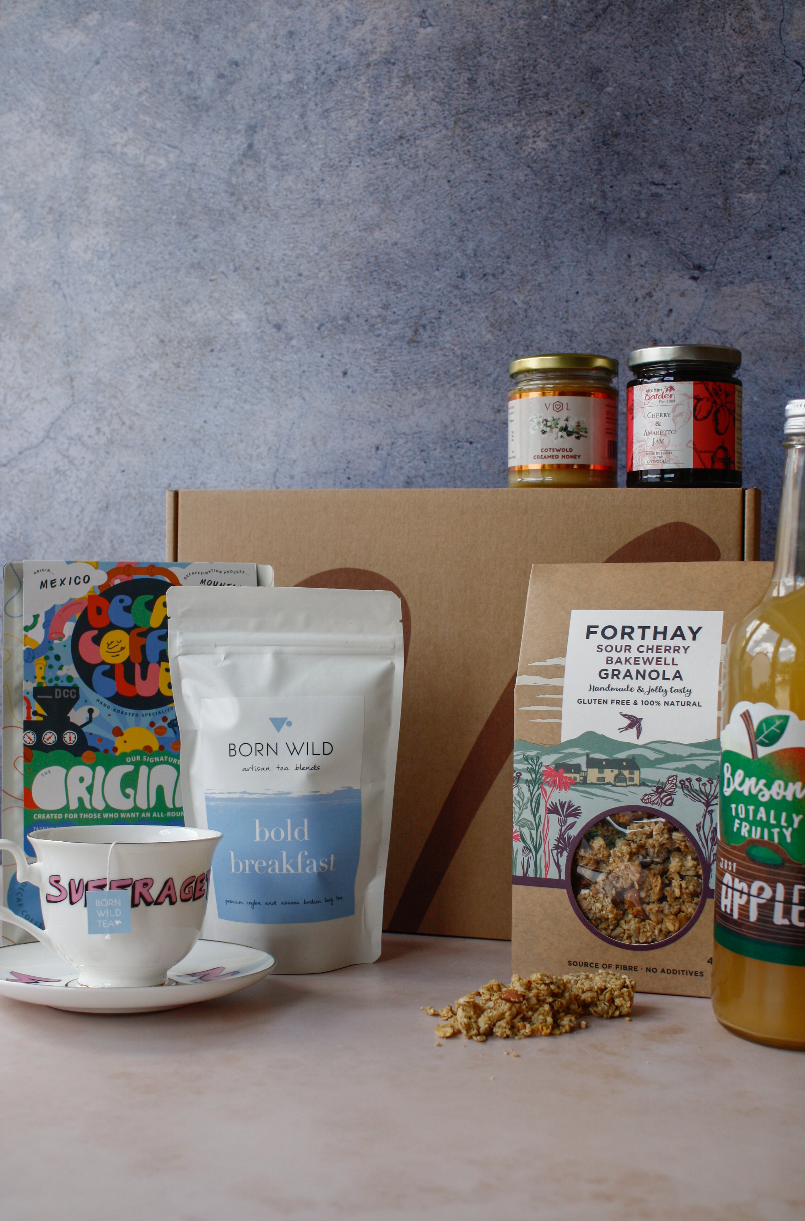 Assorted breakfast items including granola, coffee, tea, apple juice, jam and honey on a marbled surface with a gray background.