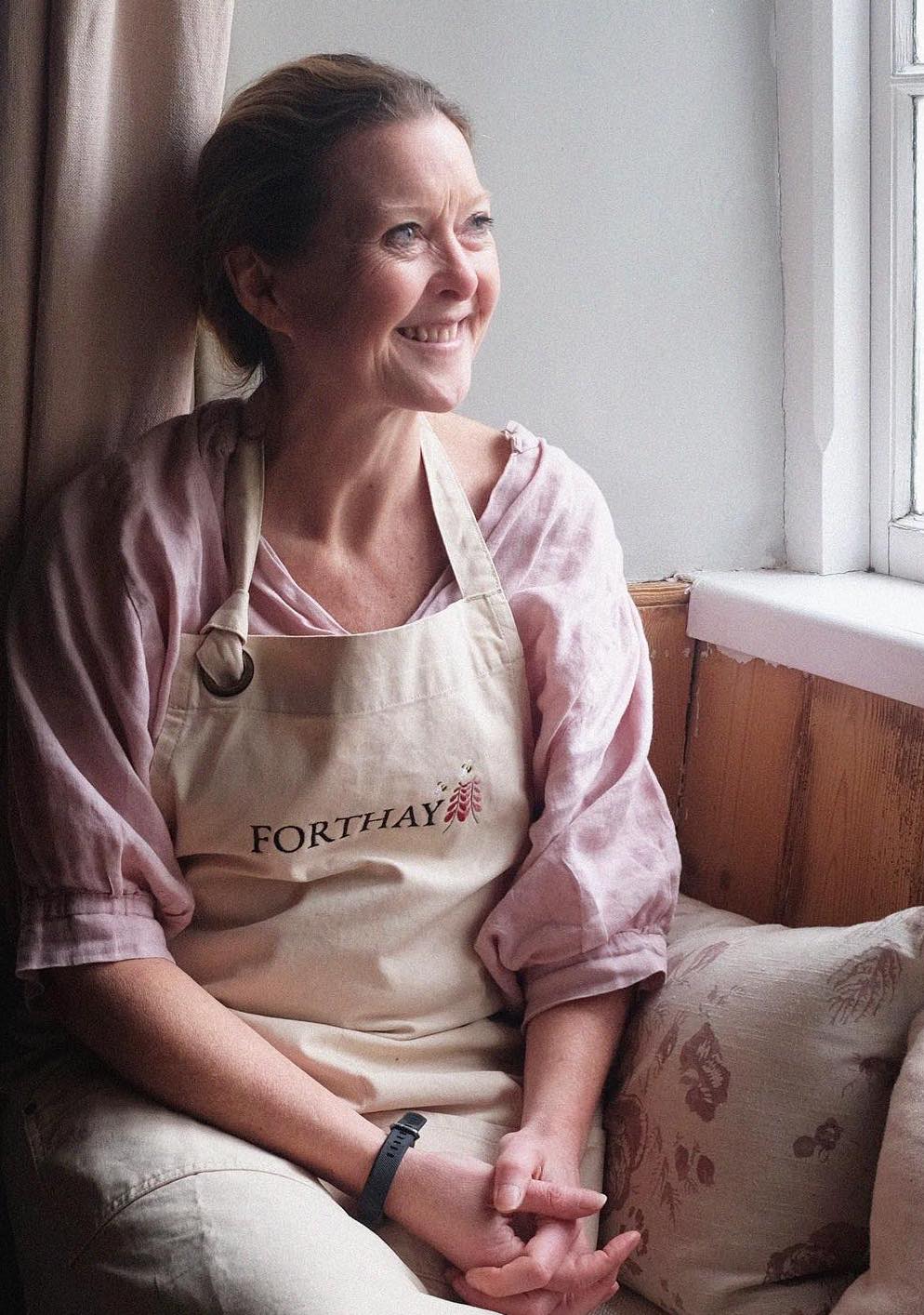 Woman wearing a 'Forthay' apron sitting by a window.