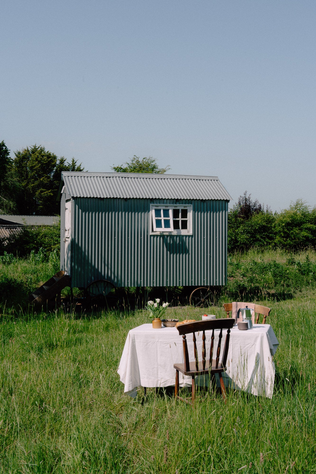 A small green corrugated metal cottage sits in a grassy field under a clear blue sky, with a small outdoor dining table and chairs set up in the foreground on the grass.