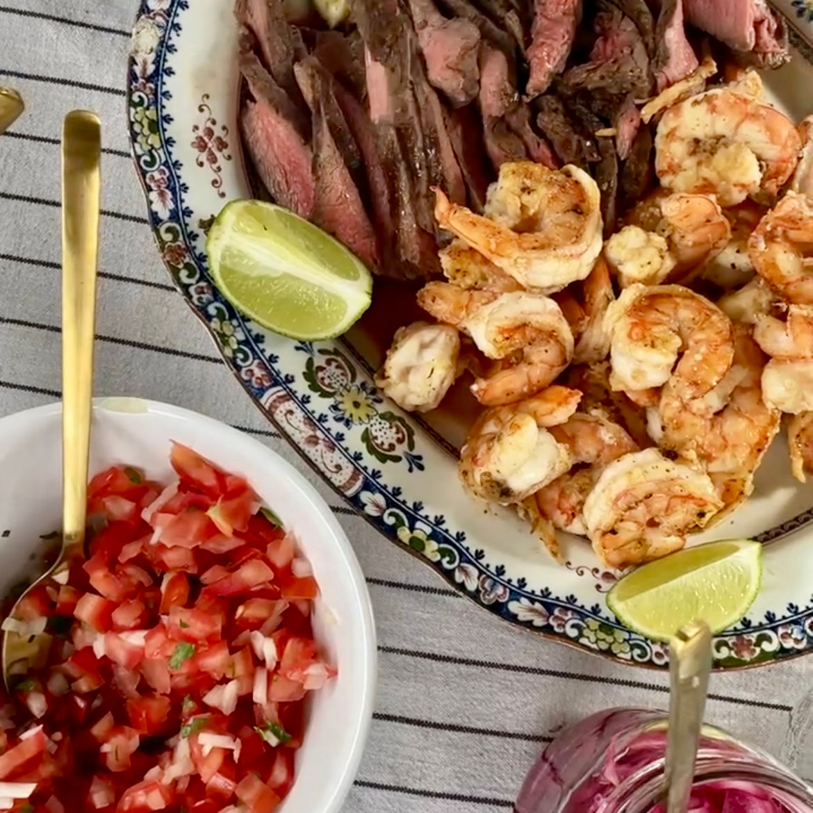 Platter of grilled meats, shrimp, and a bowl of pico de gallo on a striped tablecloth.