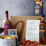Gourmet food items including a bottle of wine, crackers, cheese, and cured meats on a wooden board with a gray background.