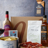 Gourmet food items including a bottle of wine, crackers, cheese, and cured meats on a wooden board with a gray background.