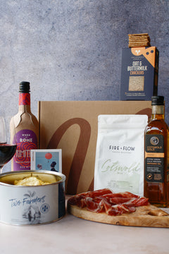 Gourmet food items including a bottle of wine, crackers, cheese, and cured meats on a wooden board with a gray background.