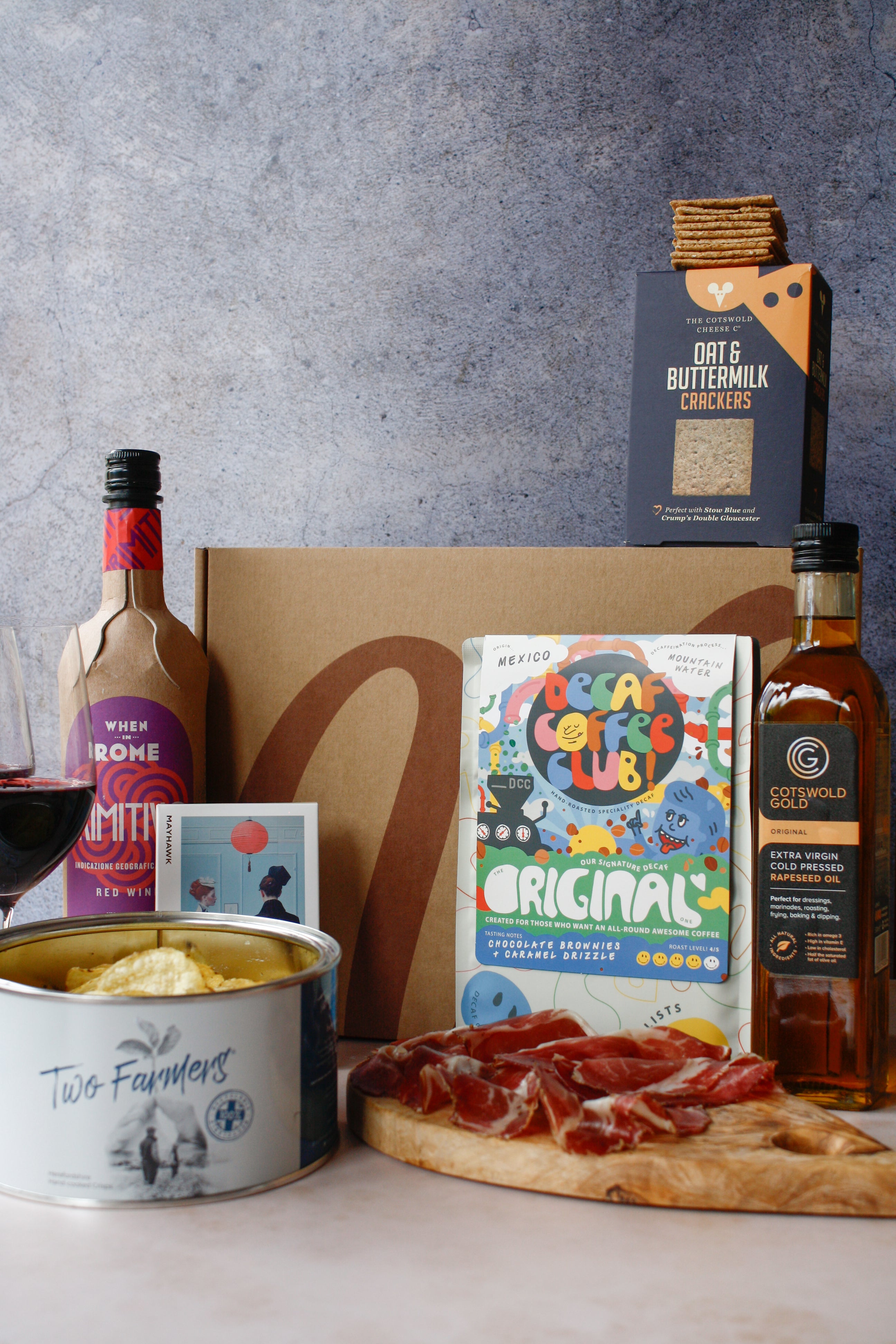 Gourmet food items including a bottle, a can, a box of crackers, and a cutting board with cured meats on a gray surface.