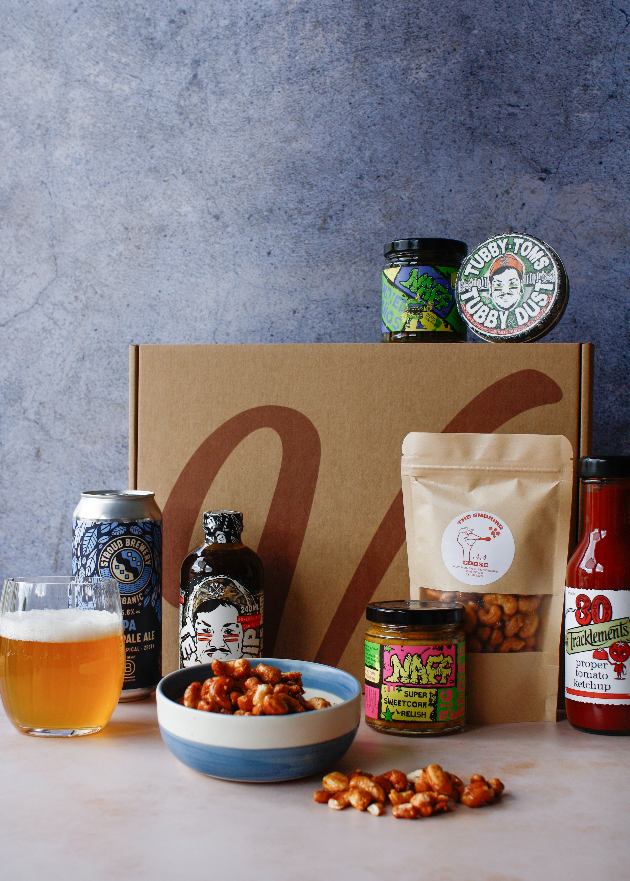 Assorted barbecue food items including a bag of nuts, a can of drink, assorted jars and spice tin on a textured surface.