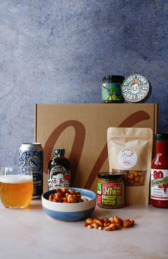 Assorted barbecue food items including a bag of nuts, a can of drink, assorted jars and spice tin on a textured surface.